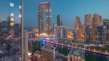 Dubai Marina gökdelenleri ve JBR bölgesi güneş doğmadan önce aydınlanmış lüks binalar ve gece gündüz geçiş zamanları ile. Palmiyeler ve kanalda yüzen teknelerle dolu bir liman.