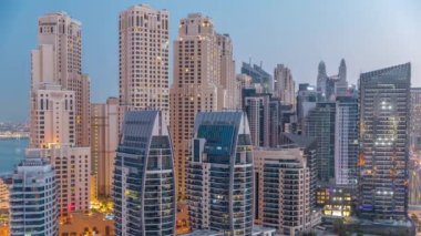 Dubai Marina gökdelenleri ve JBR bölgesi güneş doğmadan önce aydınlanmış lüks binalar ve gece gündüz geçiş zamanları ile. Palmiyeleri ve yeşil çimenleri olan limana.