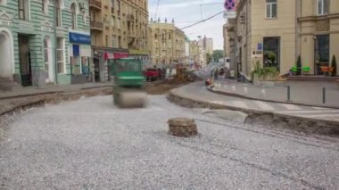 Yeşil titreşimli sıkıştırıcı yol yapımında ve zaman ayarında bir taşın üzerinde yuvarlanıyor. Arka planda kazıcı var. Tramvay raylarının yeniden inşası