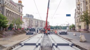 Yol inşaatı alanındaki panoramik zaman dilimine vinçle beton plakalar yerleştiriliyor. Şapkalı ve üniformalı endüstriyel işçiler. Şehir caddesindeki tramvay raylarının yeniden inşası