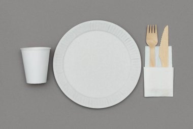 White paper dish, paper cup, wooden fork and knife in white napkin on gray background, perspective view. Set of eco friendly disposable tableware from natural material.