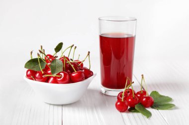 Taze kiraz suyu bardağının yanında olgun kirazlar ve beyaz ahşap tahtaların üzerinde kirazlarla aydınlık duvara yaslanmış bir kase..