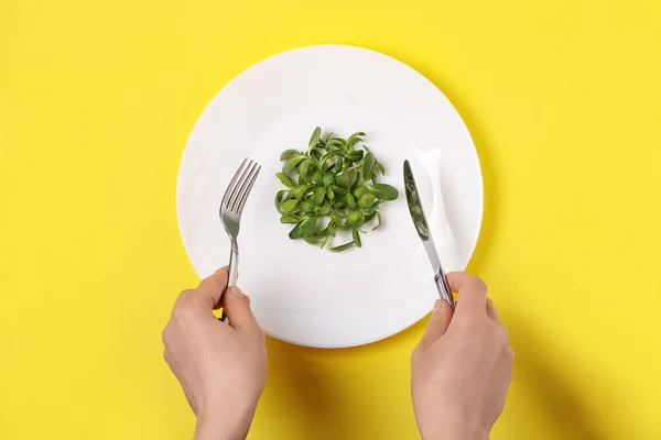 Female hands holding fork and knife over microgreens sunflower sprouts in white dish on yellow background, top view. Vegetarian food. Healthy eating concept.