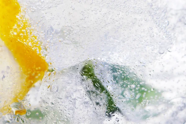 Close up of water with lemons, leaves of mint and ice cubes in glass, selective focus. Abstract background.