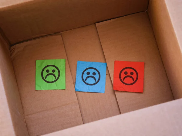 Three paper notes with sad faces on them in a cardboard box. Close up.