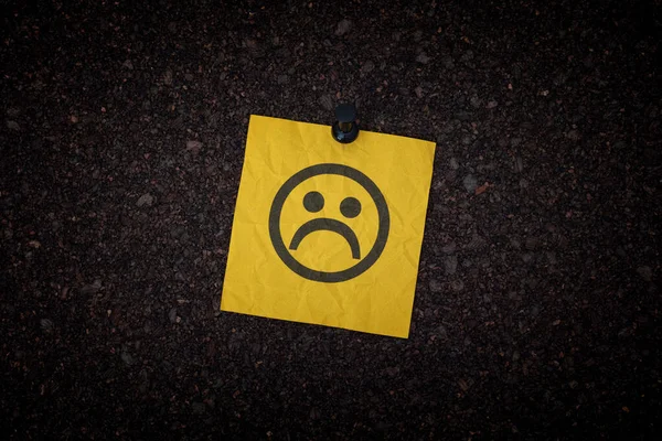 A sticky note with a sad face on it pinned to a corkboard. Close up.