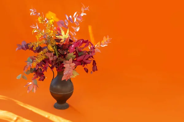 Ramo de otoño de hojas rojas y amarillas en un florero de cobre antiguo en rayos de sol sobre un fondo naranja. —  Fotos de Stock