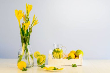 Meyve kompozisyonu ve limon parçaları, nane yaprakları, filizler, bir sürahide limon içeceği, hafif bir arkaplan karşısında doğal ışık altında alınan sarı çiçekler. Nane ve limonlu kullanışlı taze içecek..