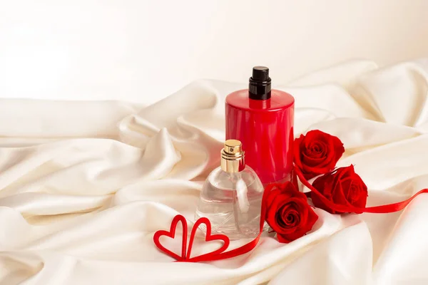 Frascos de perfume masculino y femenino, rosas rojas, corazones decorativos sobre fondo de tela satinada. Presentación de cosméticos. regalo de perfume en el día de San Valentín —  Fotos de Stock