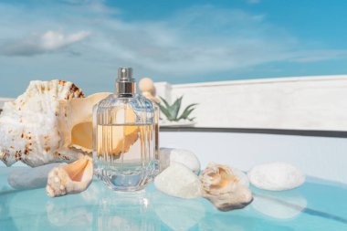 transparent spray bottle of womens or mens perfume with shells on the glass surface of the new swimming pool and tropical plants on the terrace and blue sky in summer.Presentation of womens perfume