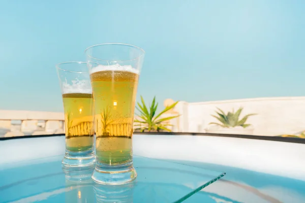 Glasses with cool beer on a glass table above the swimming pool on the terrace of a tropical hotel on a hot summer day.cooling drink at pool party.