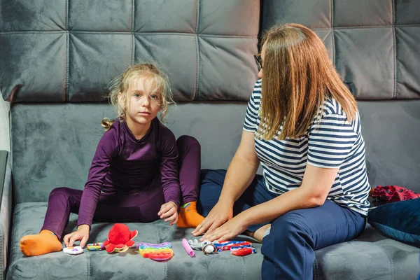 A blonde-haired girl in purple thermal underwear plays toys with her mother, sitting on the gray couch in the living room