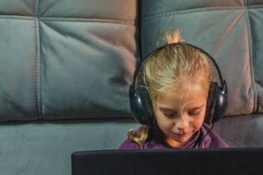 Kafasında büyük kulaklıklar olan küçük bir kız internette öğrenmek için dizüstü bilgisayar kullanıyor. Uzaktan eğitim