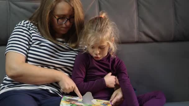 眼鏡をかけた女性は 灰色のソファの上で家に座っている子供に絵本を読んでいる母親です 幸せな家庭の概念 ロシアのクラスノダール2021年9月25日 — ストック動画