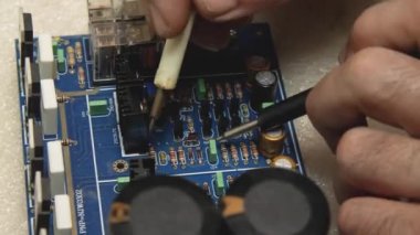 Usta tamirci mavi elektronik panoda bir testçiyle pin teşhisi koyuyor. Elektronik, bilgisayar, telefon ve televizyon tamirhanesi.