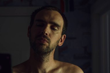 Tişörtsüz tıraşsız beyaz bir adam akıllı telefona bakıyor.