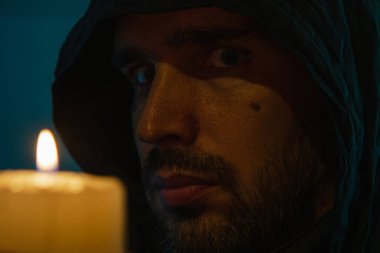 Beyaz, traşsız, siyah kapüşonlu bir adamın yüzünü mum alevinin arka planına yakın çekim. Yan görünüm