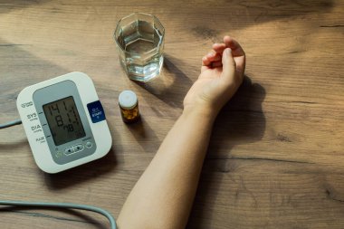 Masanın üzerinde bir bardak su, bir şişe hap ve bir otomatik tonometre var. Bir kadının eli masanın üzerinde duruyor. Cihazın ekranında yüksek tansiyon görünüyor. Sağlık Konsepti