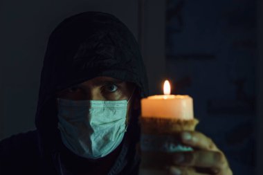 Elinde yanan bir mumla siyah kapüşonlu ve tıbbi maskeli beyaz bir adamın yüzü. Salgında keder kavramı