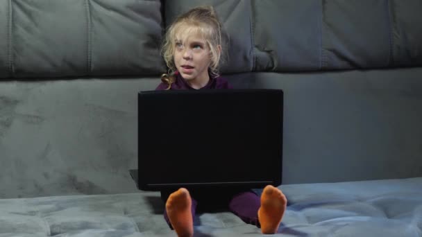En flicka barn med en bärbar dator sitter på en grå soffa och ler. Begreppet lycklig barndom. — Stockvideo