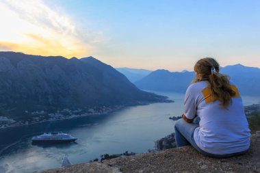 Deniz koyundaki bir yolcu gemisine bakan bir kız. Dağın arka planında ve günbatımı gökyüzünde. Karadağ 'da turizm kavramı