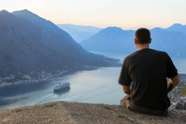 Siyah tişörtlü bir adam deniz kıyısındaki bir yolcu gemisine bakıyor. Bir dağın arka planına karşı. Gün batımından önce. Karadağ 'da turizm kavramı