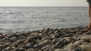 Bir kadının çıplak ayaklarının kayalık bir plajda yürürken görüntüsü. Arka planda su sıçrıyor.