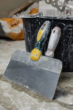 Siyah plastik bir kovaya yaslanmış beton zeminde duran iki mala. Arka planda boş kağıt alçı torbaları var. Oda yenileme konsepti