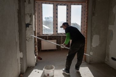 Şapkalı bir adam elinde alüminyum bir kuralla boş bir odada duruyor. Bir dekoratör odayı alçılıyor. Oda yenileme konsepti