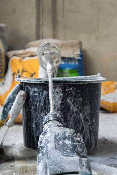 Kuru mikser başlığıyla delik delen kirli siyah bir kova. Arka planda beton bir duvar ve kağıt alçı torbaları var. Mülkün onarım ve dekorasyon konsepti