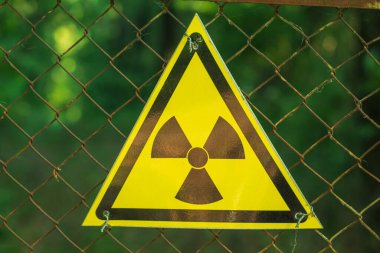 Bir tel örgüdeki sarı üçgen radyasyon işaretinin yakın çekimi. Arka planda yeşil bir orman var. Tehlike uyarısı