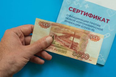 Adamın elinde Covid-19 'dan aşı sertifikası almak için para var. Mavi arka plan. Yakın plan. Çeviri: yeni bir koronavirüs enfeksiyonuna karşı aşı sertifikası COVID-19