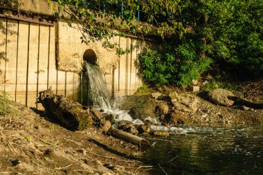 Kirli kanalizasyon suyu beton duvardaki bir borudan nehre akıyor. Suyun yüzeyinde beyaz köpük vardır, arka planda yeşil yapraklı ağaçlar vardır. Ekoloji kavramı