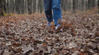 Sonbahar ormanında yaprakların üzerinde yürüyen beyaz spor ayakkabılı ve kot pantolonlu kadınlar. Düşen yaprakları saçan kadınların yavaş çekim görüntüleri. Yakın plan. Kamerada hareket