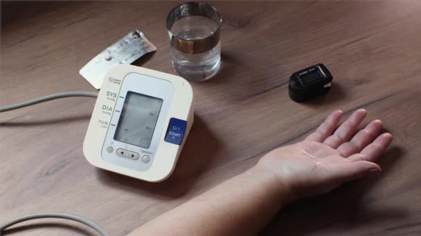 一位老年妇女用自动血压监测器测量她的血压 在一张木制桌子上有一只手 一个装置 一杯水和一些药丸 2021年9月12日 俄罗斯克拉斯诺达尔 — 图库视频影像