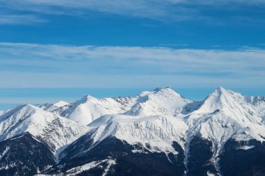 Mavi gökyüzü ve bulutlardan oluşan bir arka planda karlı dağ zirveleri. Dağlar aşağıdan siyahtır, ortası ve tepeleri tamamen beyazdır. Güzel bir kış manzarası