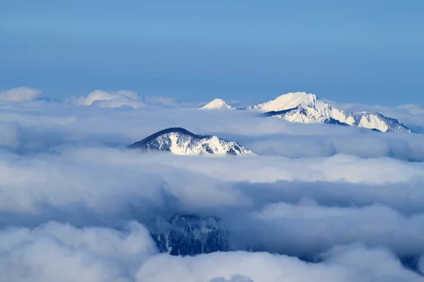雪の山のピークがふわふわの雲の外に固執する 背景には澄んだ青空が広がっています 冬の風景 — ストック写真
