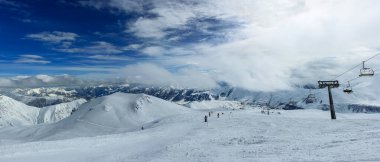Sandalyesi olan bir kayak merkezi. Piste ve karlı dağların bulutlarla örtülü görüntüsü. Bulutların arasından mavi gökyüzünü görebilirsin. Panoramik fotoğraf