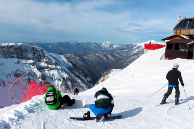 Kayak pistinin tepesinde snowboardcular kaykaylarını takıyor ve kayakçılar yoldalar. Arka planda dağ sıraları, mavi gökyüzü ve bulutlar var. Tatillerde kış sporları