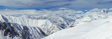 Tepeleri bulutlarla örtülü karlı dağlar. Bulutların arasından mavi gökyüzünü görebilirsin. Panoramik fotoğraf
