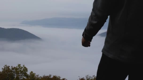 Un hombre camina hasta el borde del acantilado, mira a su alrededor y levanta la mano. Hay montañas en la niebla frente a él. Un estilo de vida activo. — Vídeos de Stock