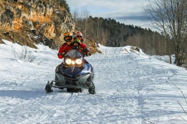 A family rides a snowmobile with lights on. In the background are rocks, woods and cloudy skies. Father child and mother on a snowmobile in the mountains. Russia, Krasnodar Territory, January 3, 2011 clipart