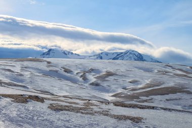 Kış dağı manzarası. Karla kaplı dağ zirveleri karla kaplı dağların üzerinde yükselir. Arka planda mavi bir gökyüzü ve tüylü bulutlar var.