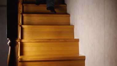 Kot pantolon ve siyah çoraplı bir adam ikinci kattan ahşap bir merdivenden iner. Akşam, evde loş bir ışık yanıyor.