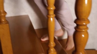 Kamera, gri pantolonlu bir kadınla yan yana hareket ediyor. Bacaklar ahşap bir merdivene tırmanıyor. Evde loş bir ışık yanıyor. Yavaş çekim.