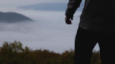 Pantolon ve sweatshirt giyen bir adam uçurumun kenarına doğru yürür ve elini kaldırır. Önünde dumanla kaplı güzel bir dağ geçidi var. Aktif bir yaşam tarzı.