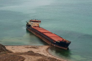 Gemi enkazı. Eski, paslı bir kargo gemisi kıyıda karaya oturmuş. Petrol ürünlerinin izleri suda görülebilir. Terk edilmiş gemi