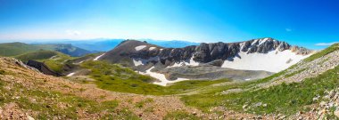 Panorama of a mountain landscape. View of the mountain range with the remnants of last year's snow. Blue sky in the background. Rocks and grass in the foreground