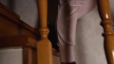 Slow motion, the camera follows a woman's legs in gray pants and black socks walking down a wooden staircase. View from the side over the balusters. The light in the house is dim