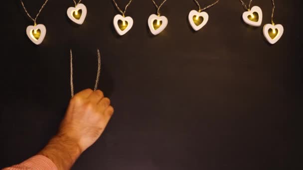 A mans hand writes in white chalk on a black board I love you. A garland of hearts hangs in the background. Valentines Day holiday concept. — Stock Video